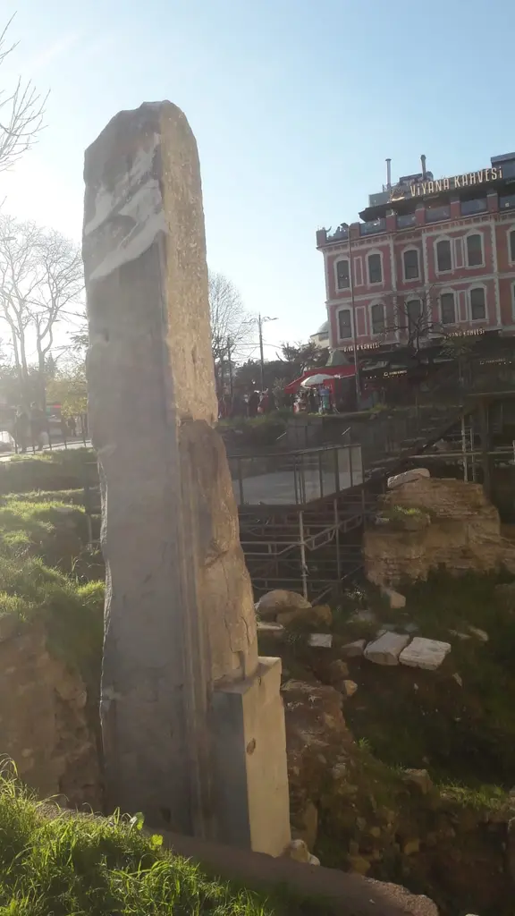 Güneş karşısında Milyon Taşı; aşınmış mermer ayak, arkada kazı alanının taş kalıntıları, iskeleler ve kırmızı cepheli eski bir bina.