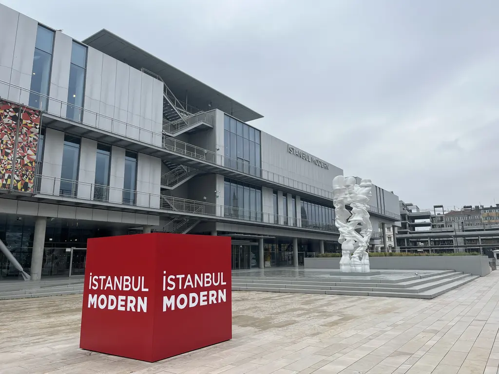 İstanbul Modern'in önünde üzerinde müzenin adı yazan kırmızı büyük küp; arkada gri cepheli bina ve beyaz bir heykel.