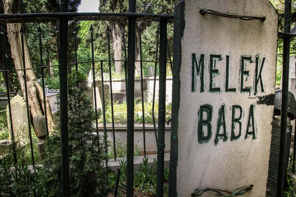 Karacaahmet'te Melek Baba kabri; demir parmaklık, üzerine yeşil harflerle "Melek Baba" yazılmış taş ve arkada ağaçlar.