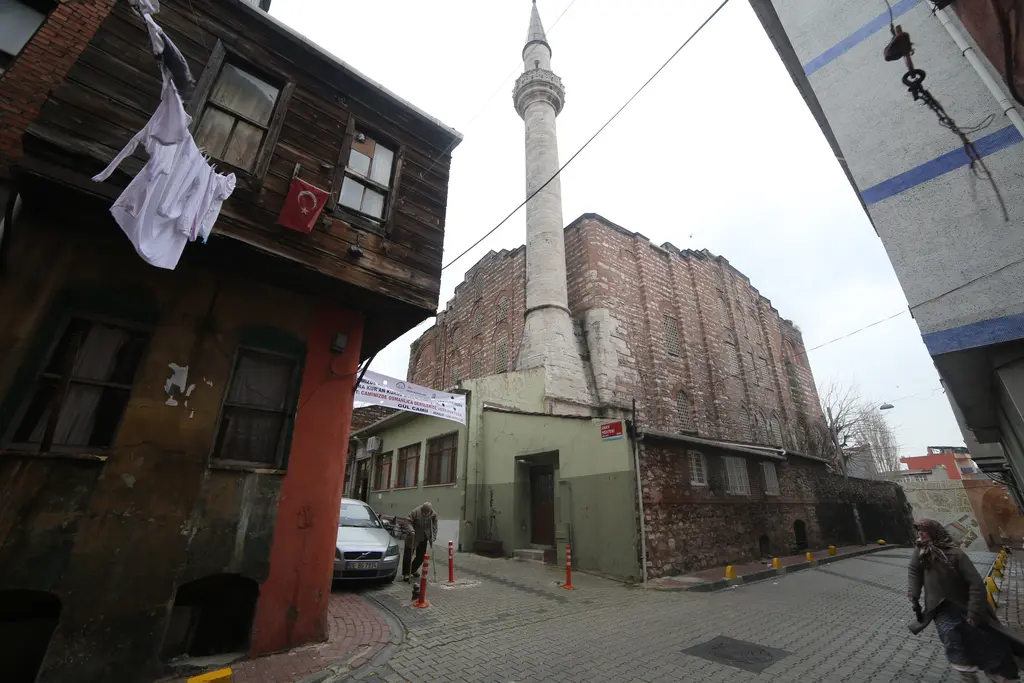 Gül Camii'nin bulunduğu sokak; solda cumbalı eski ahşap ev, sağda caminin taş ve tuğla yan duvarı ile minare, kışın çıplak ağaçlar ve iki yaya.