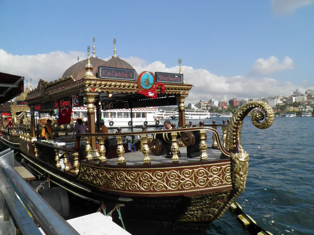 Eminönü rıhtımında yaldızlı süslü tekne; kubbeli çatılar, kıvrık pruva, bayraklar ve arkada Haliç ile karşı kıyı.