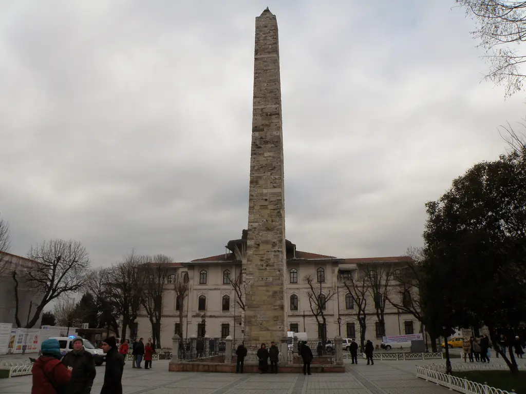 Bulutlu bir günde Örme Sütun; önde insanlar, sağda koyu yeşil ağaçlar, arkada iki katlı taş bina ve meydan.