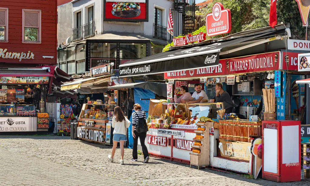Ortaköy'de kumpir tezgâhları; renkli malzeme kapları, tabelalar, sıra bekleyenler ve arkada eski evler.