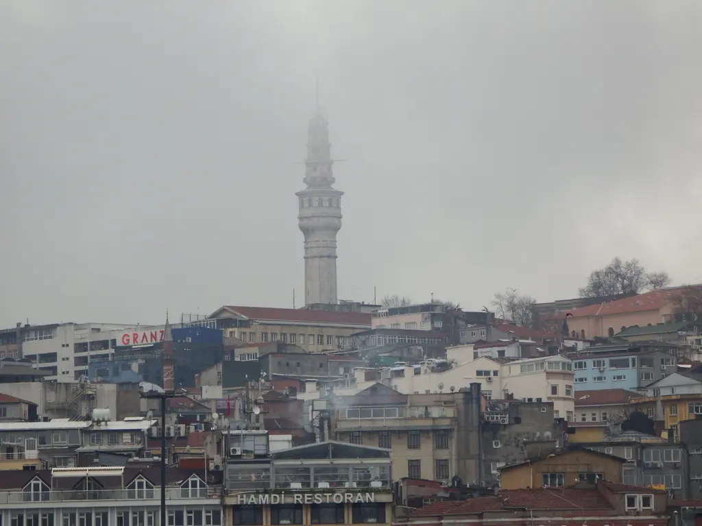 Eminönü yönünden Beyazıt Kulesi; önde yoğun kent dokusu ve çatılar, puslu havada uzakta yükselen kulenin ince silueti.