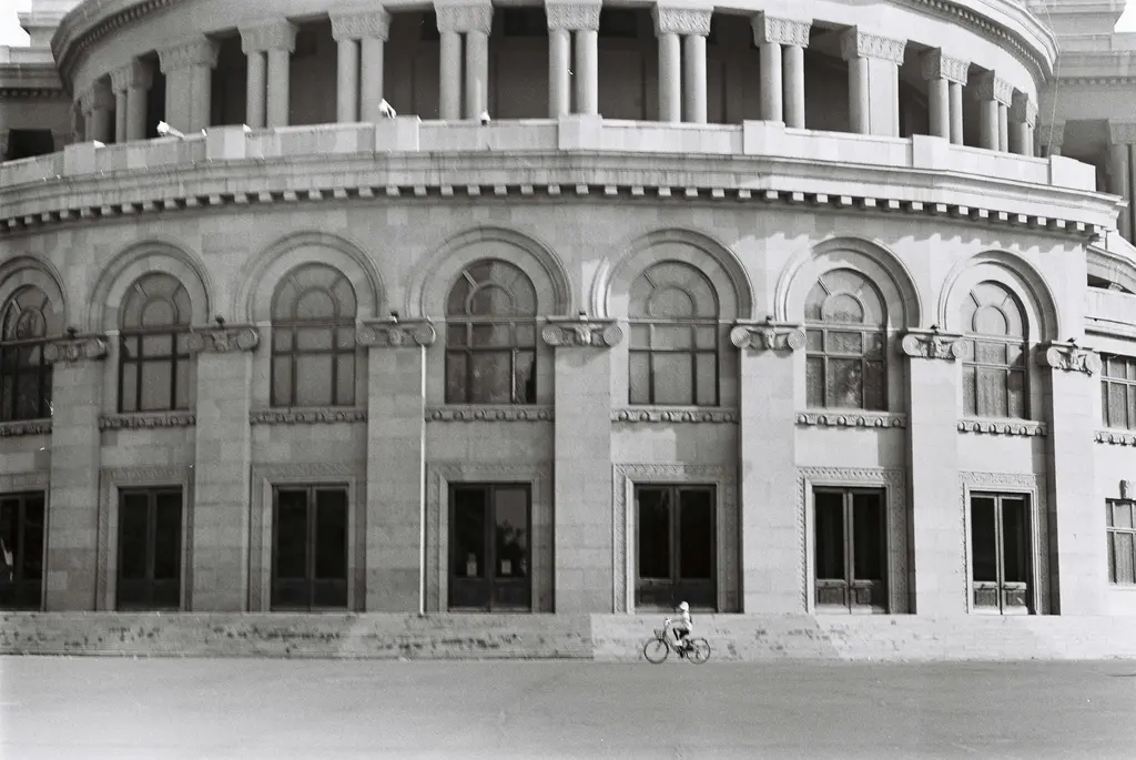 Erivan Opera ve Bale Tiyatrosu'nun yuvarlak taş cephesi siyah-beyaz bir karede; üst katta çepeçevre sıralanan sütunlar, altta kemerli pencere dizisi ve binanın önünden geçen bisikletli.