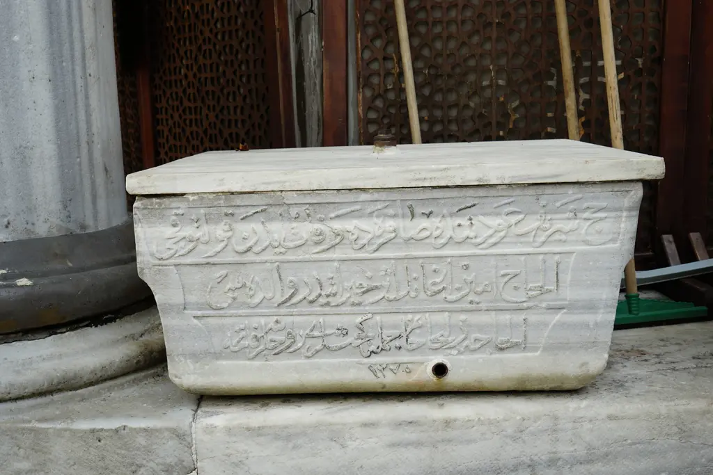 Yeni Valide Camii'nin avlusunda su teknesi; üzerinde Osmanlıca yazı kazılı mermer tekne ve ahşap kafes.