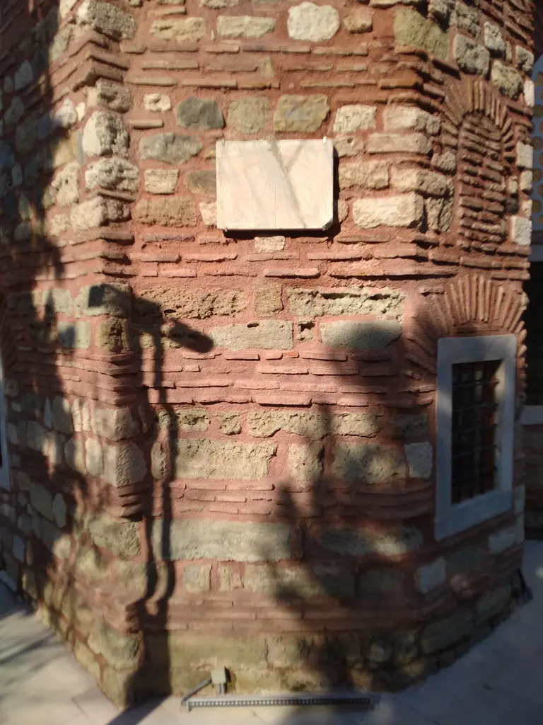 Atik Mustafa Paşa Camii'nin köşesinde duvara gömülü kare mermer levha; almaşık tuğla ve taş örgü, sağda demir parmaklıklı pencere, duvara düşen ağaç gölgeleri.
