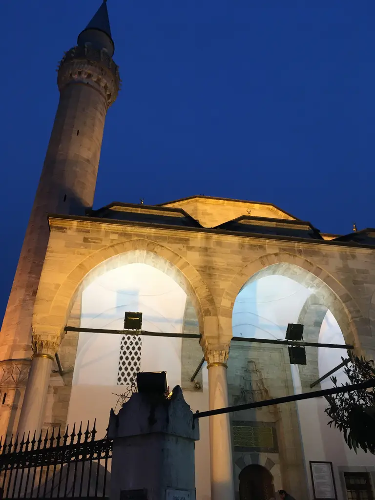 Gece Firuz Ağa Camii'nin aydınlatılmış son cemaat revakı; sivri kemerler, mermer sütunlar, arkada yükselen minare ve koyu mavi gökyüzü.