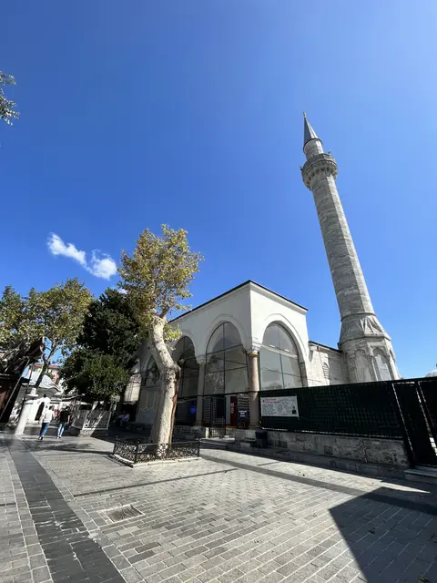 Fatih'te Koca Mustafa Paşa Camii'nin ön cephesi; camekânla kapatılmış üç kemerli son cemaat revağı, beyaz taş gövde, sağda tek şerefeli minare ve önde gri parke meydan.