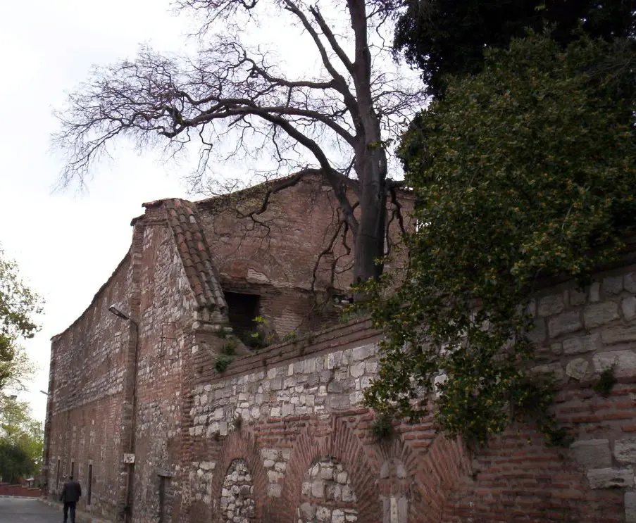 İmrahor İlyas Bey Camii kışın; çıplak ağacın arkasında almaşık örgülü uzun duvar ile yıkık üst kotlar, sağda sarmaşık, önde kaldırımda yürüyen bir kişi.