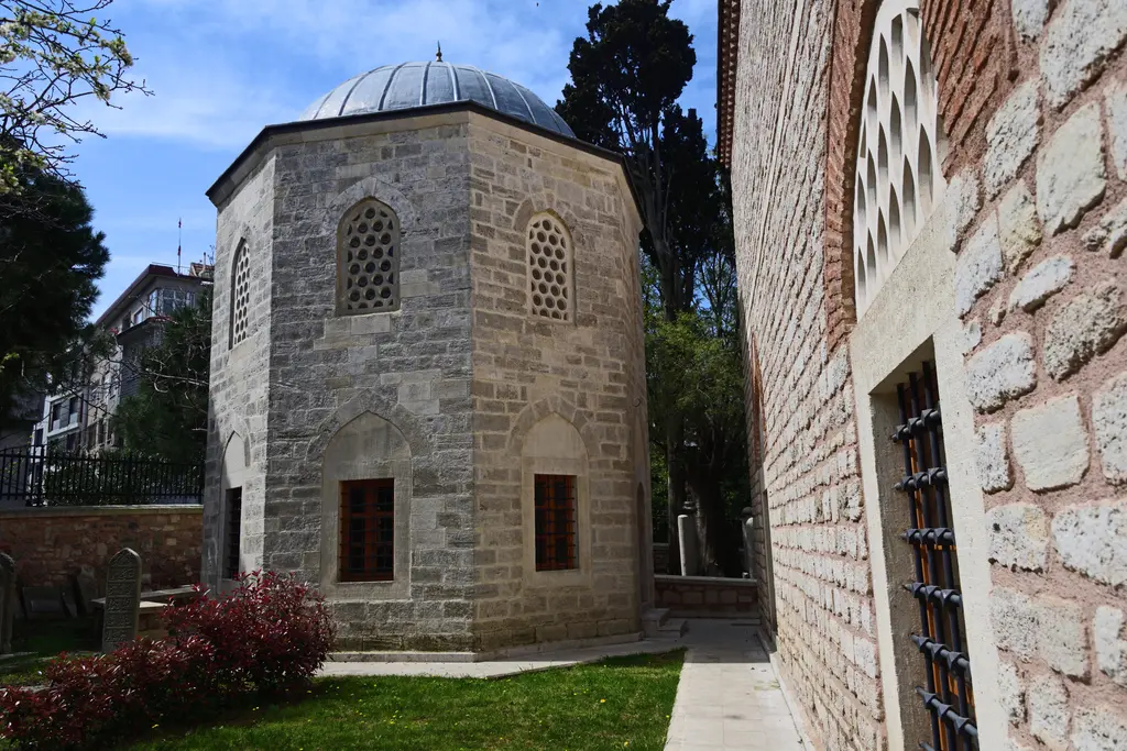 Rum Mehmed Paşa'nın türbesi; sekizgen taş gövde, kafesli üst pencereler, kırmızı çiçekli çalılar ve sağda cami duvarı.