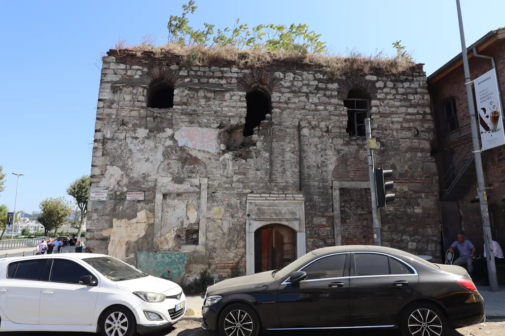 Ayakapı Hamamı'nın girişi; sıvası dökülmüş taş cephe, kemerli kapı, önde park etmiş beyaz ve siyah arabalar.
