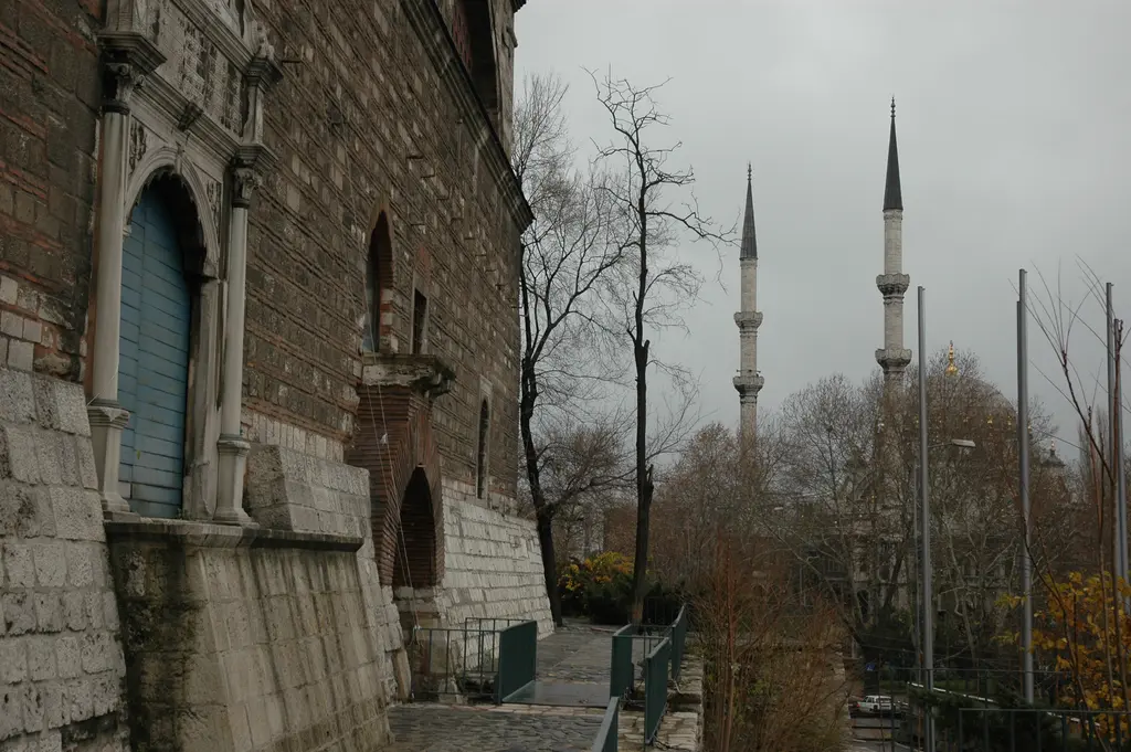 Tophane-i Âmire'nin yeşil boyalı kapısı; mermer çerçeve, yanında taş duvar ve uzakta Nusretiye Camii'nin ince minareleri.