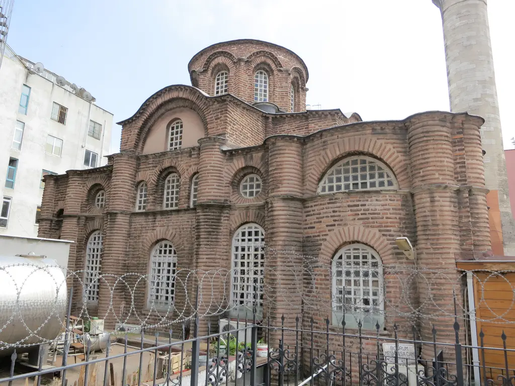 Bodrum Camii'nin kuzey cephesi kapalı havada; payandalar, kemerli kafesli pencereler, arkada kubbe kasnağı, önde jiletli tel sarılı parmaklık ve cami levhası.