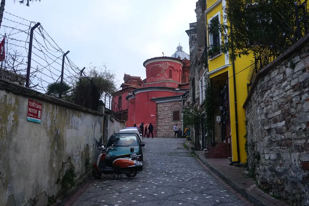 Fener'de yokuşlu arnavut kaldırımı sokak; solda dikenli telli yüksek duvar, sağda sarı bir ev ve yokuşun sonunda Kanlı Kilise'nin kırmızı kubbe kulesi.