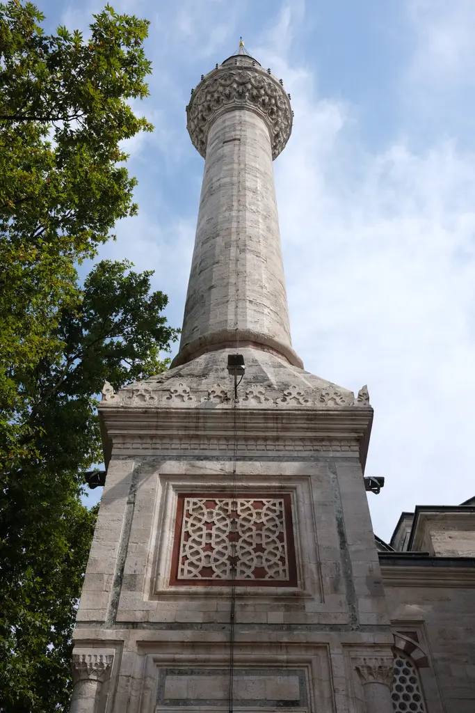 Beyazıt Camii'nin minaresi aşağıdan; taş gövde, tek şerefe, şerefe altındaki mukarnas çıkmalar, kurşun külah ve önündeki ağaç dalları.