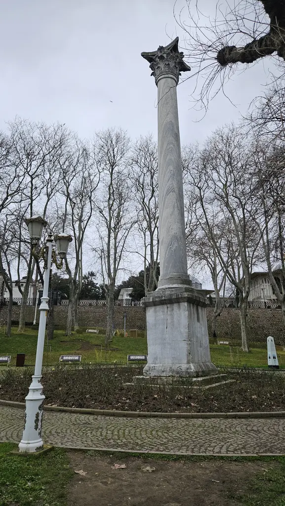 Kışın Gotlar Sütunu; toprak tarhın ortasında kaide ve gövde, önde beyaz bir lamba direği, arkada yapraksız ağaçlar ve surlar.