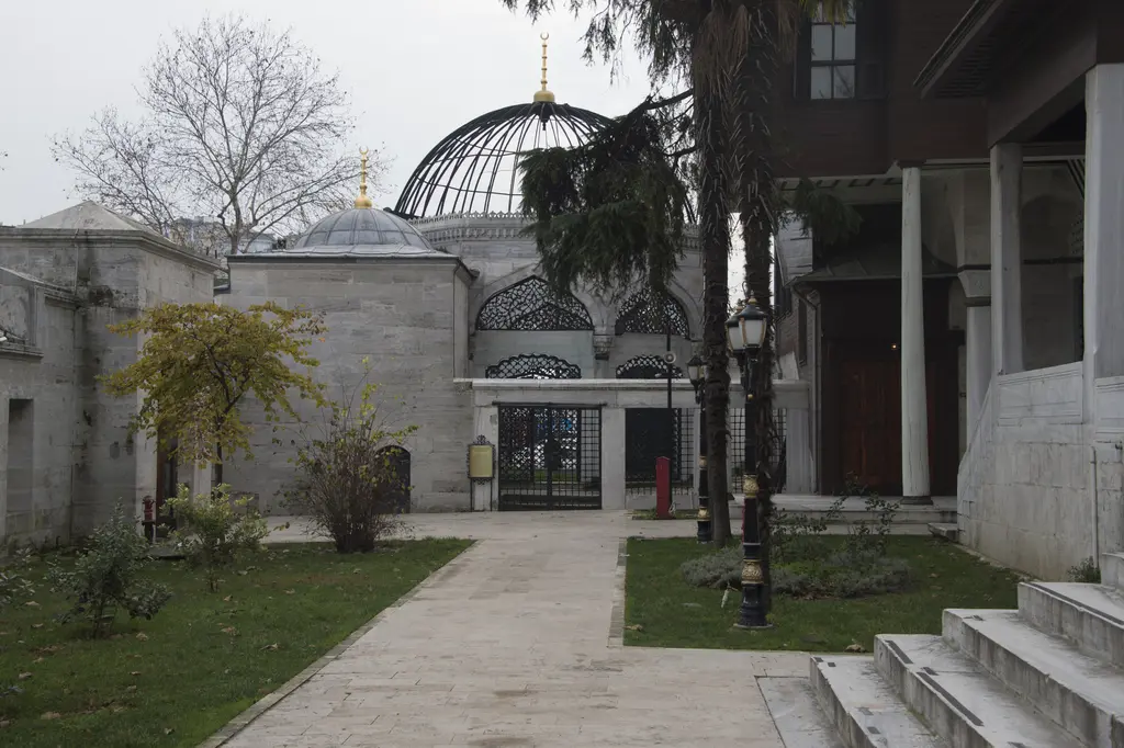 Yeni Valide Camii'nin dış avlusu; ağaçlar, taş yol, merdiven ve arkada demir kafes kubbeli türbe.