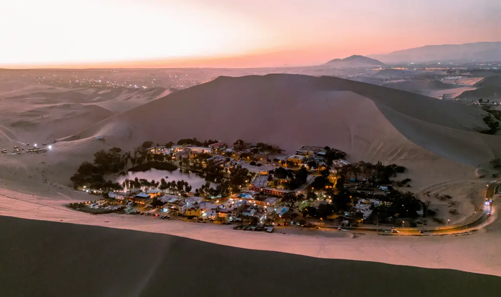 Huacachina vahası; gün batımında kum tepelerinin ortasında uzanan göl, çevresini saran palmiyeler ve ışıkları yanan kasaba.