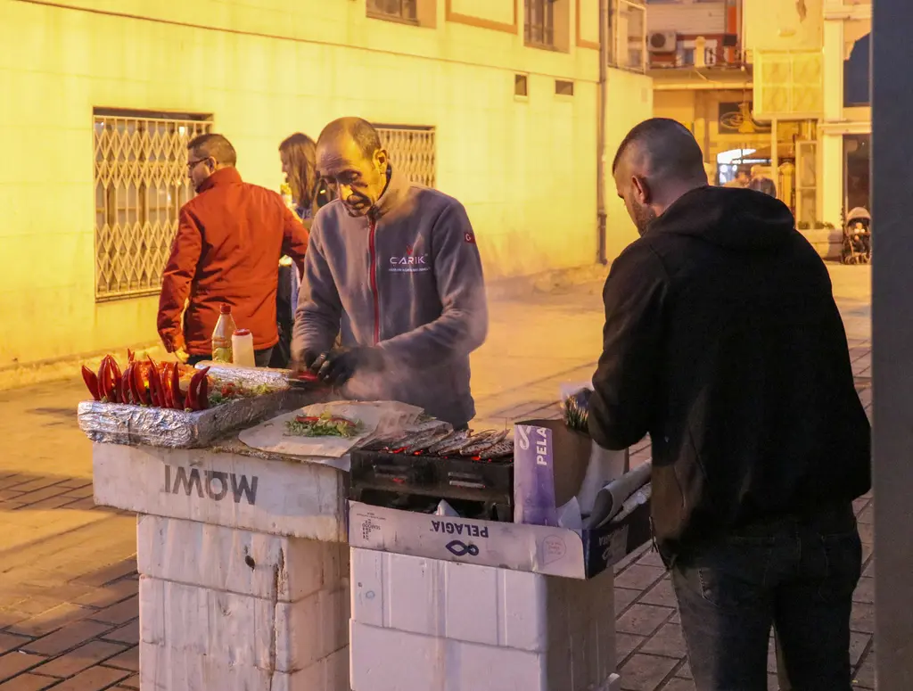 Karaköy'de akşam balık dürüm yapan satıcılar; ızgaradaki balıklar, tezgâhta ekmekler ve sarı ışıkta bir duvar.