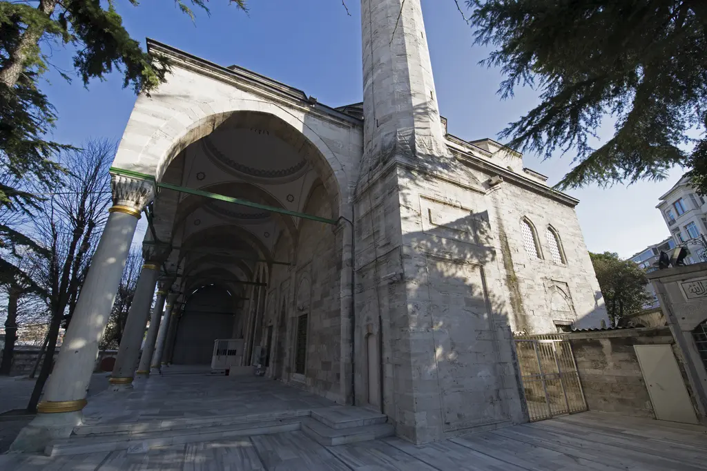 Gazi Atik Ali Paşa Camii'nin revakı yandan; sivri kemerli kubbeli bölüm, sütunlar, kesme taş duvar, yükselen minare ve gölgeler.