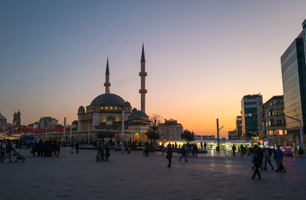 Taksim Meydanı'nda alacakaranlık; turuncu ufkun önünde caminin kubbesi ve minareleri, meydanda yürüyen insanlar ve sağda cam cepheli bina.