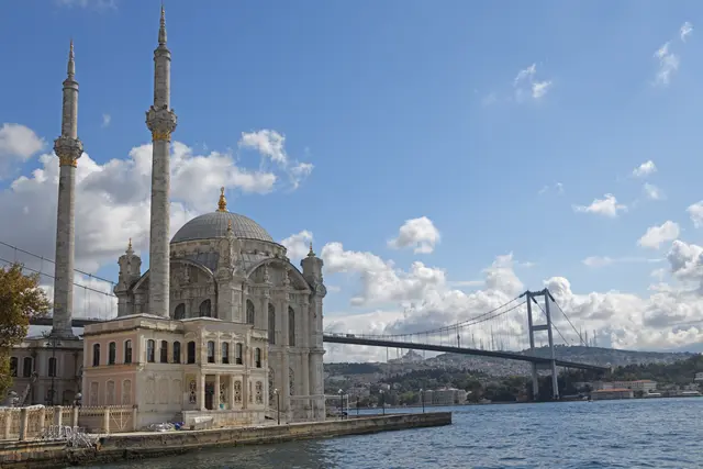 Beşiktaş'ta Ortaköy Camii; kıyıdaki beyaz taş cami, iki ince minare, arkada Boğaz'ı aşan asma köprü ve bulutlu mavi gökyüzü.
