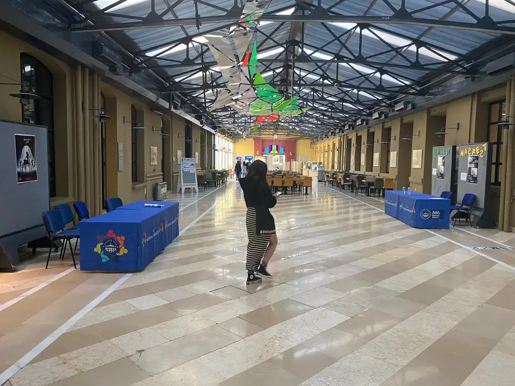 Kadir Has Üniversitesi'nin cam çatılı avlusu; iki yanda fabrika cepheleri, metal çatı makası, açık renkli taş zemin ve masalar.