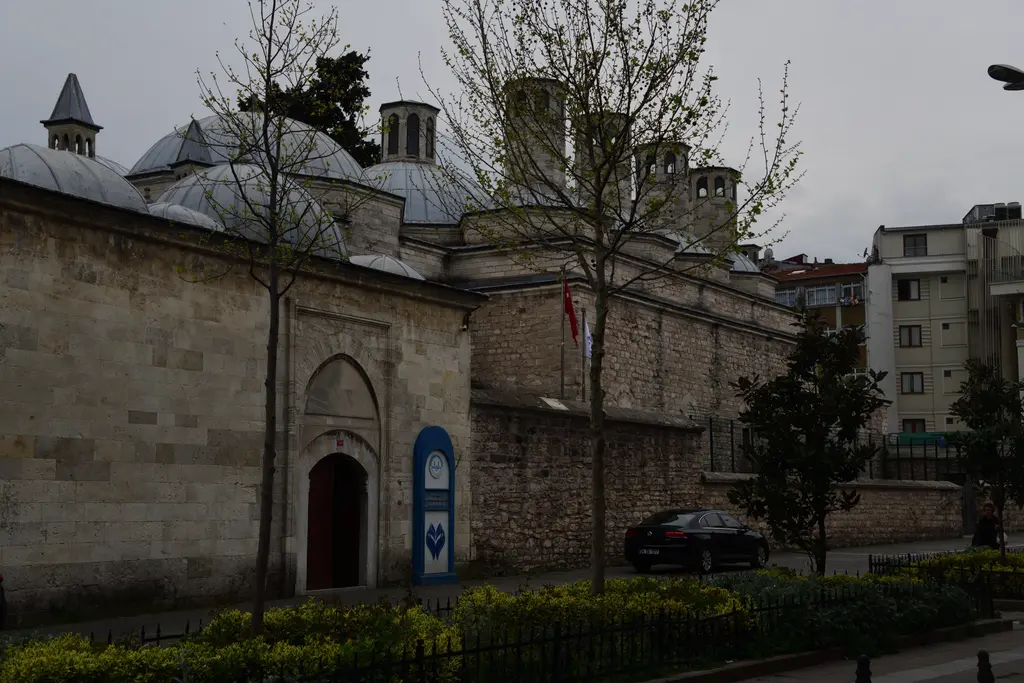 İlkbaharda Haseki Sultan Külliyesi; kubbeler ve bacalar, sivri kemerli bir kapı, yeni yapraklanmış ağaçlar ve çiçekli bahçe.