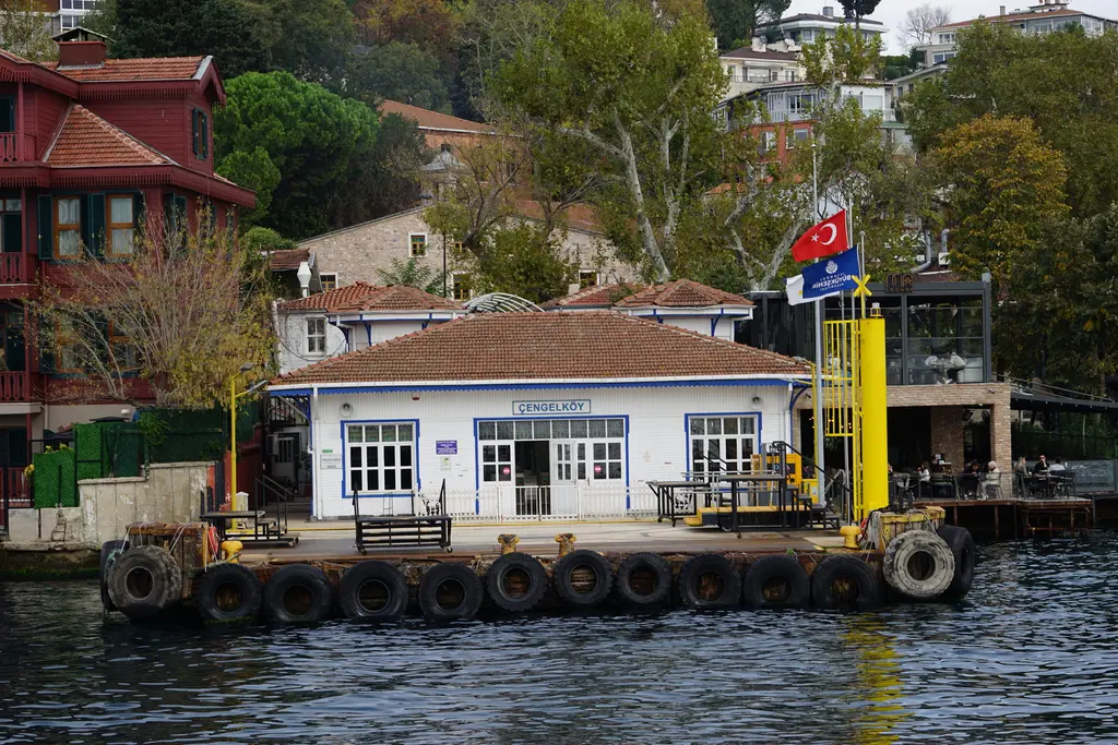 Çengelköy İskelesi; beyaz cepheli tek katlı bina, mavi pencere çerçeveleri, Türk bayrağı ve lastik kaplı rıhtım.