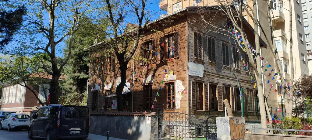 Göztepe'de Ömerpaşa Caddesi'nde köşk; renkli flamalarla süslenmiş eski ahşap yapı, ağaçlar ve yanında apartman.