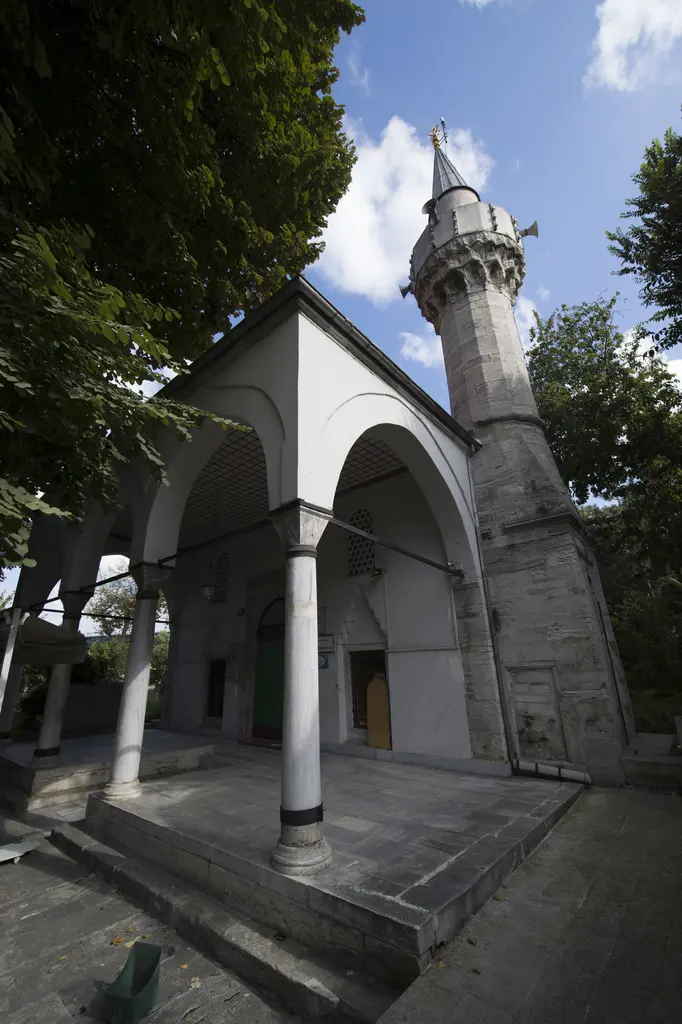Defterdar Camii'nin son cemaat yeri; iki kemerli revak, beyaz sütunlar, ince taş minare ve ağaçlar arasında mavi gökyüzü.