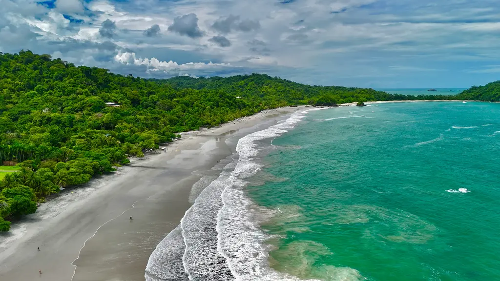 Manuel Antonio'da yukarıdan görünen kıyı; ormanla kaplı burnun eteğinde açık renkli kumsal, kıyıya paralel ilerleyen köpüklü dalgalar ve turkuazdan laciverde açılan deniz.