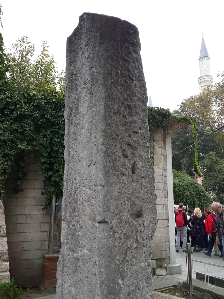Milyon Taşı'nın gri mermer ayağı; arkasında sarmaşıkla örtülü duvar, sağda kaldırımda yürüyen ziyaretçiler ve uzakta bir minare.
