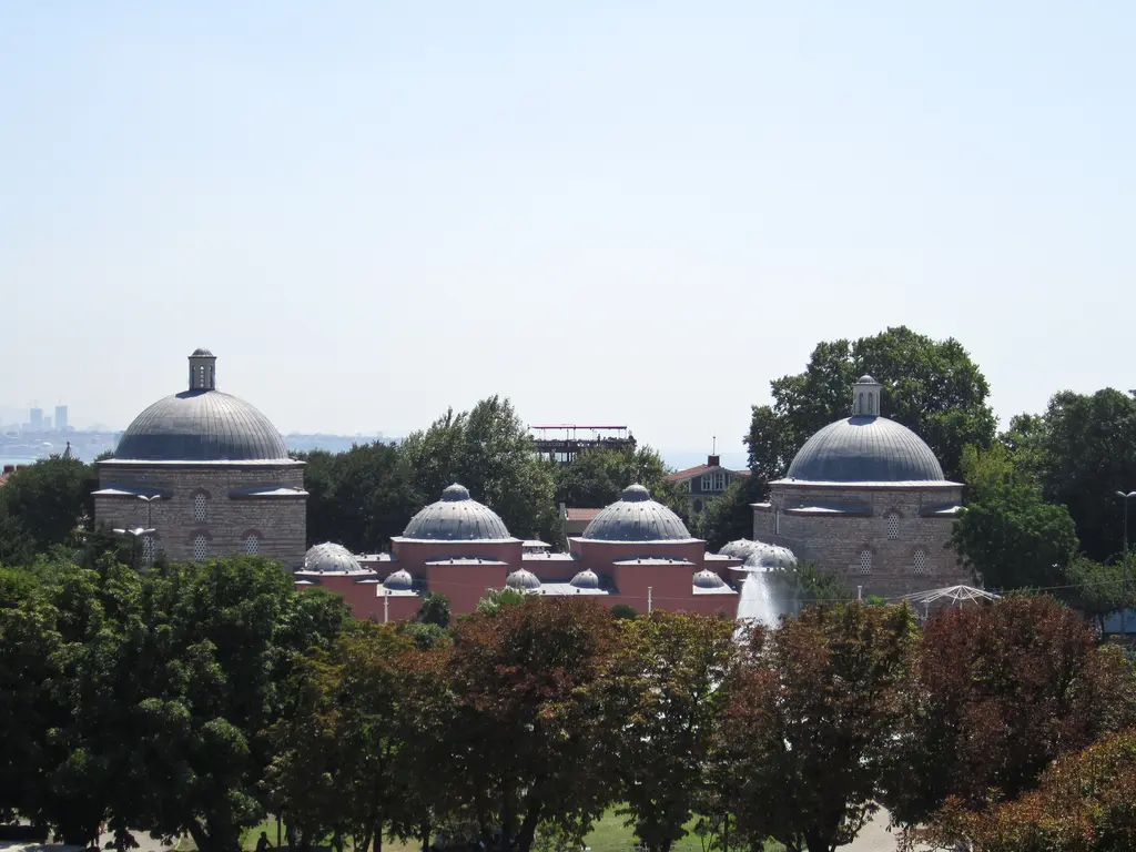 Ağaçların ardından hamamın kubbeleri; ortada büyük kubbe, iki yanında küçük kubbeler ve önde park ağaçlarının tepeleri.