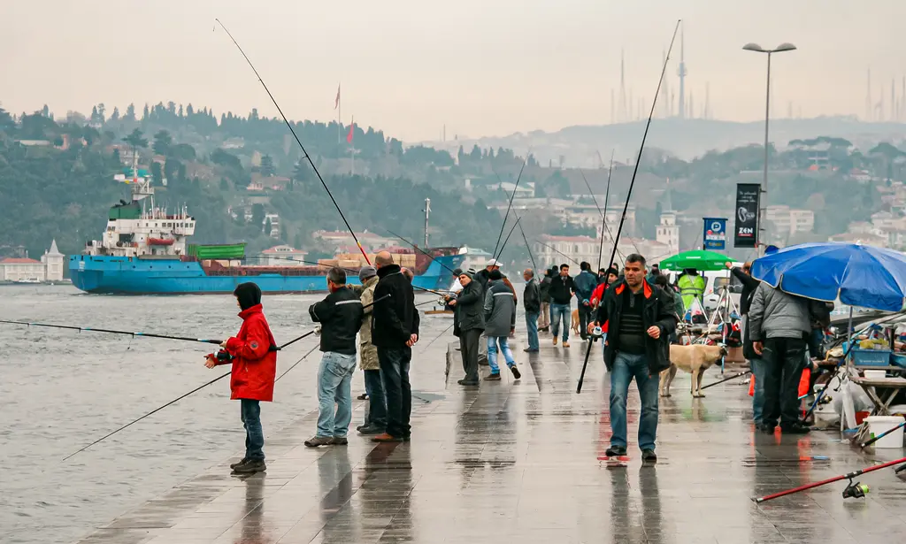 Bebek'te yağmurlu bir günde kıyıda olta atan balıkçılar; ıslak kaldırım, şemsiyeler ve arkada geçen gemi.