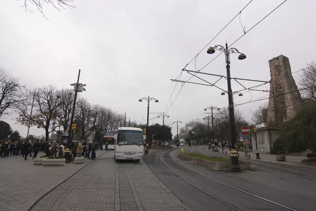 Bulutlu bir günde tramvay yolu; sağda su terazisinin kulesi, solda kalabalık kaldırım, ortada bir minibüs ve sokak lambaları.