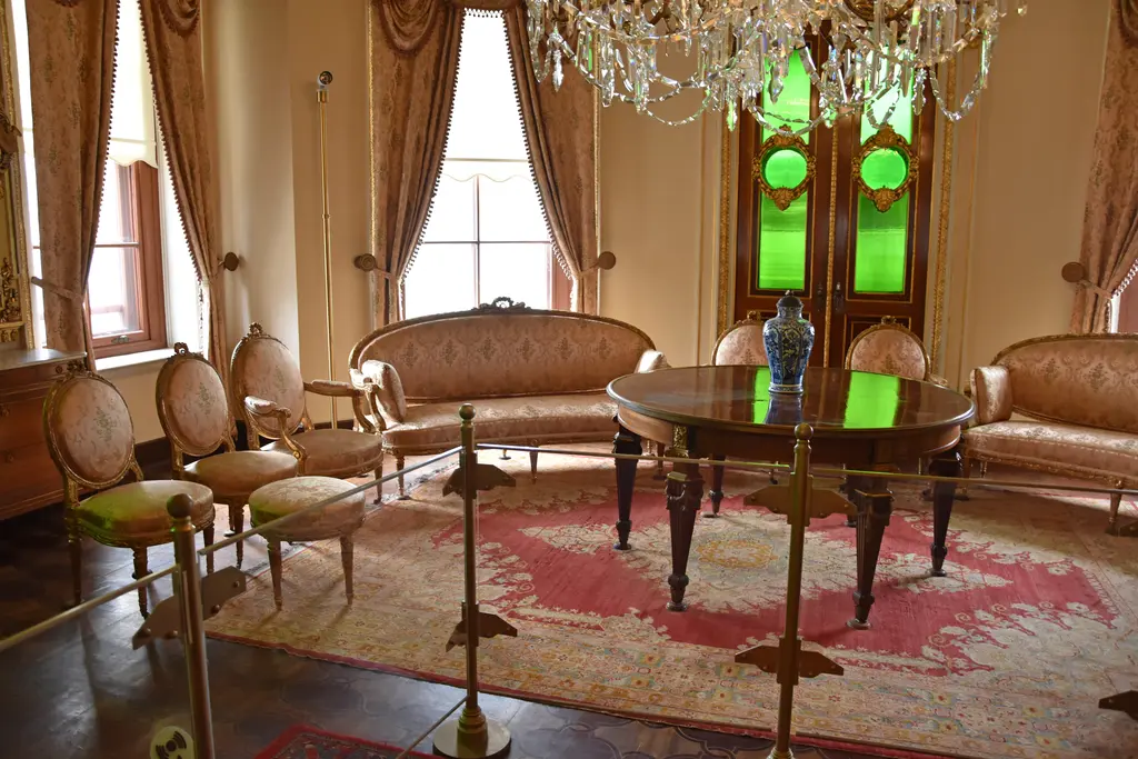 Küçüksu Kasrı'nda salon; ortada oval ahşap masa, çevresinde kumaş kaplı sandalyeler, kanepe, halı ve yeşil camlı kapı.