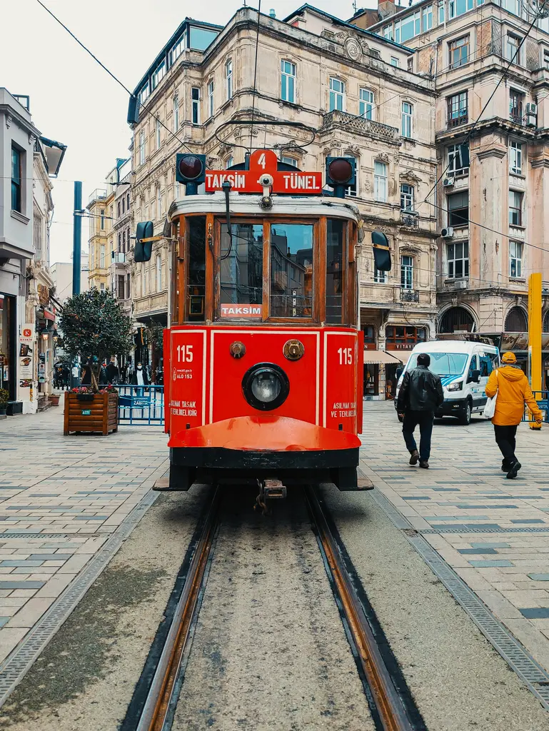 İstiklal Caddesi'nde önden kırmızı tramvay; üstünde Taksim Tünel tabelası, arkada süslü cepheli eski apartmanlar ve taş döşeli zemin.