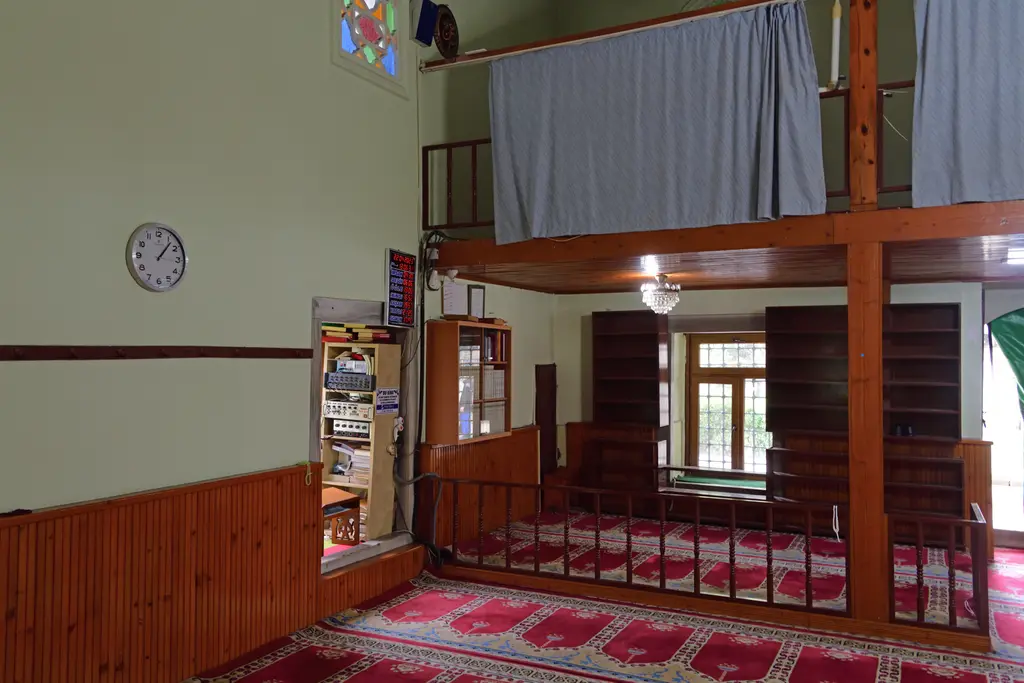 Defterdar Camii'nde mahfil; ahşap direkli üst kat, perdeler, kırmızı halı, duvar saati ve kitaplıklar.