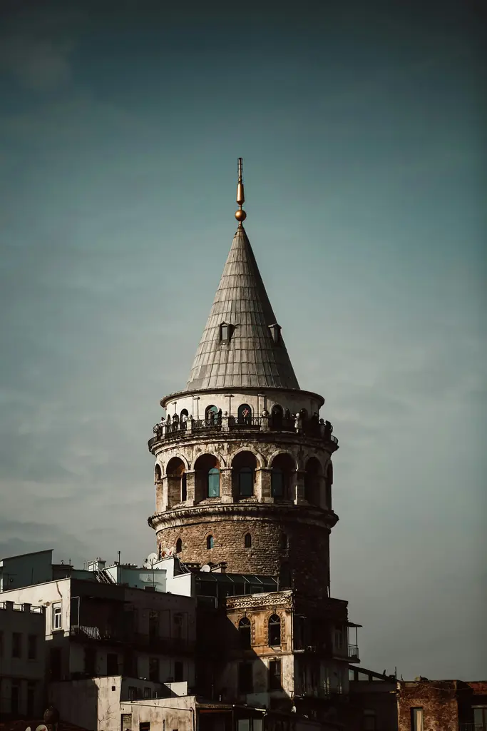 Alacakaranlıkta Galata Kulesi; koyu gökyüzüne yükselen sivri külah, kemerli pencereli üst kat ve önde gölgede kalan binalar.