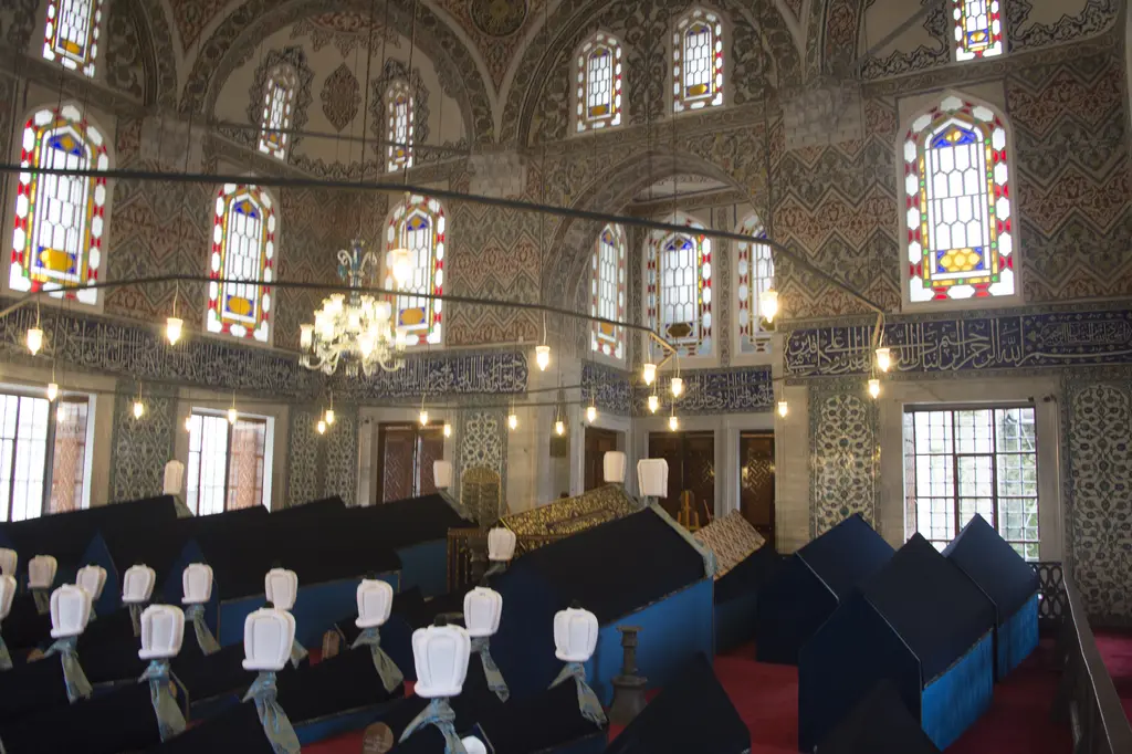 Sultan Ahmed Türbesi'nde sanduka sıraları; koyu mavi kadife örtüler, başlarında beyaz sarıklar, arkada pencereler ve çini kaplı duvar.