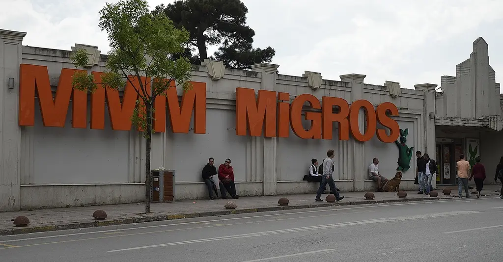 Caddebostan'da eski Migros binası; uzun alçak cephe, turuncu harflerle MMM Migros yazısı, ağaçlar ve kaldırımda oturanlar.