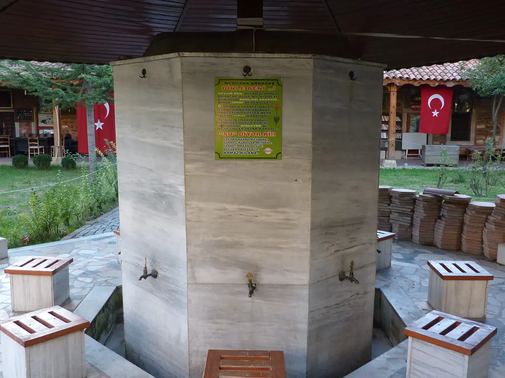 Medrese avlusunda altıgen mermer şadırvan; pirinç musluklar, sığ havuz, çevresinde ahşap taburelerle katlanmış seccadeler ve arkada Türk bayrakları asılı ahşap hücre cepheleri.