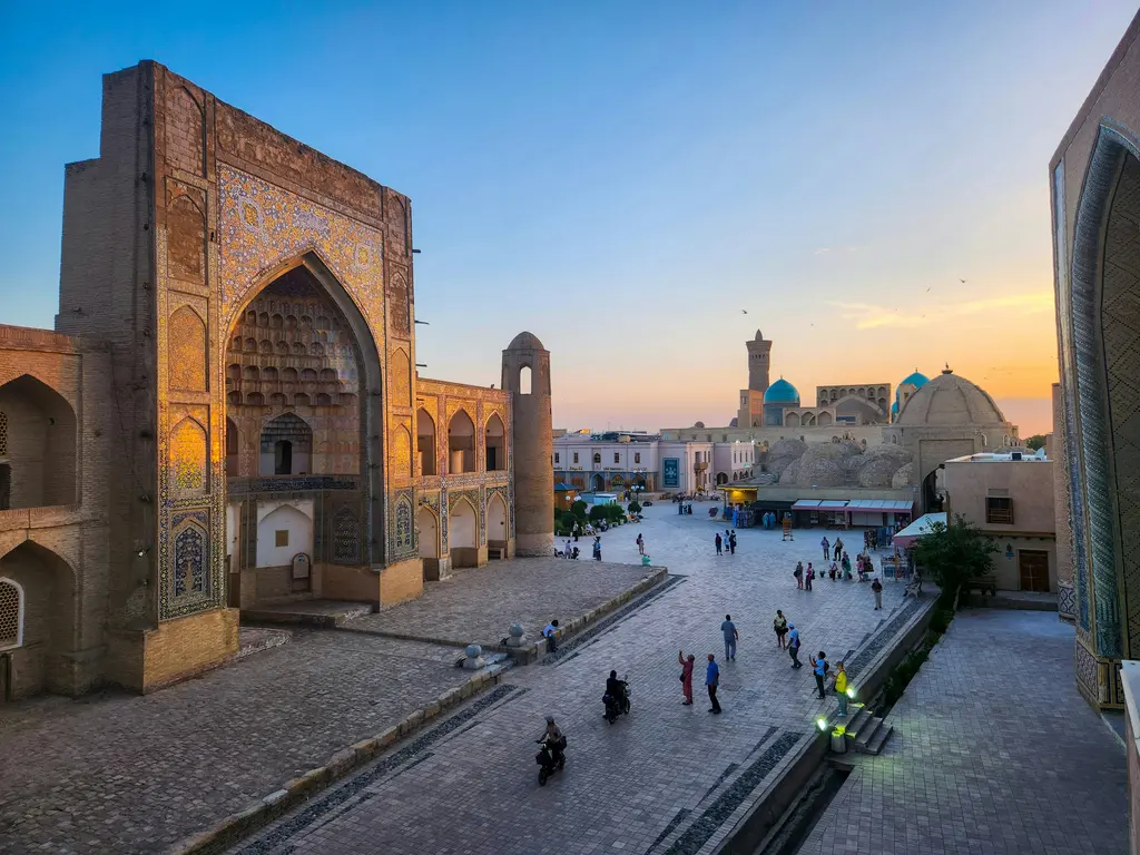 Buhara'da gün batımında Poi Kalyan meydanı; öndeki anıtsal çini portal, arkada Kalyan Minaresi ve mavi kubbeler, taş zeminde gezenler.