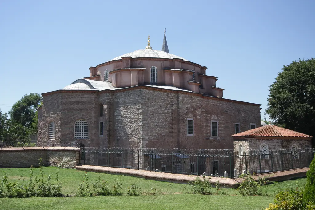Küçük Ayasofya Camii'nin güneydoğudan görünümü; kırmızı tuğla ile beyaz taşın almaşık dizildiği duvarlar, alçak kurşun kubbe ile altın alem, solda minare ve önde geniş yeşil çim.