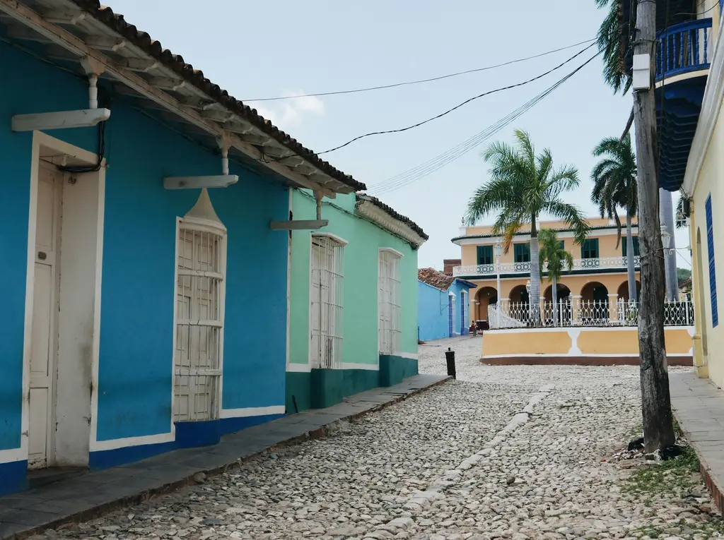 Küba'nın UNESCO korumasındaki Trinidad kasabasında arnavut kaldırımlı sokak; mavi ve yeşil sömürge evleri, palmiyeler ve arkada sarı bir konak.