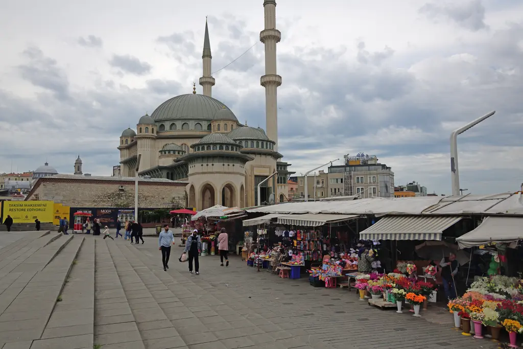 Taksim'de çiçek tezgâhları ve Taksim Camii; önde tenteli tezgâhlar ve saksılar, arkada kubbeler ve minare.
