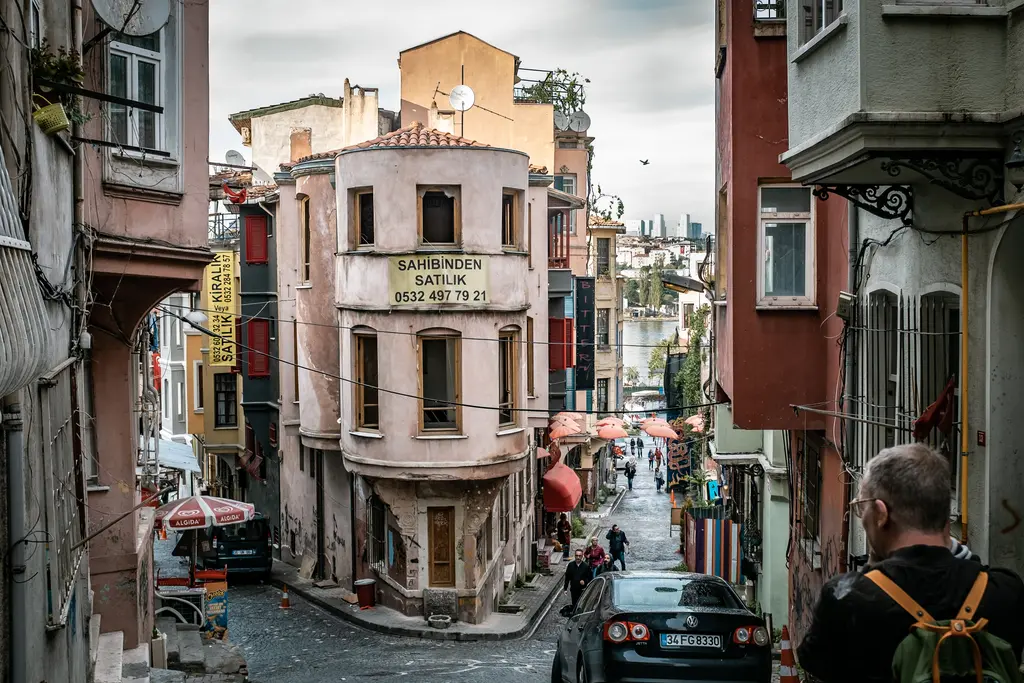 Balat'ta sokak kavşağında köşe evi; pembe cepheli yüksek bina, satılık tabelası, iki yanda cumbalı evler ve yayalar.