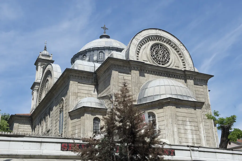 Aya Triada Kilisesi'nin doğu tarafı; yarım kubbeler, kubbe kasnağı, çan kulesi, önde bahçe ağaçları ve mavi gökyüzü.